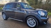 A dark-colored MINI compact SUV is parked on a muddy, rural path surrounded by leafless trees and bushes. The vehicle’s body is glossy, reflecting the bright sunlight filtering through the cloudy sky. The SUV features five doors, silver alloy wheels, and chrome side mirrors. The ground is wet, suggesting recent rain.