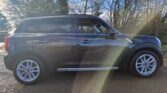 A dark grey MINI SUV is parked on a road with a backdrop of leafless trees and a clear sky. Sunlight creates a lens flare on the vehicle's side, and the car's windows are tinted. The sporty silver alloy wheels shine against the dry road surface.