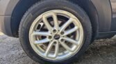 Close-up of a car's wheel with a silver alloy rim featuring five double spokes and a central cap displaying the brand "MINI." The tire is fitted on a dark-colored vehicle parked on a gravel surface. The tread pattern and sidewall markings are clearly visible.