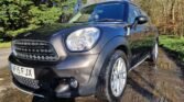 A black MINI Countryman car is parked on a wet asphalt road surrounded by trees. The car's windows are slightly tinted, and the license plate reads "WF15 FFJX." The reflection of the MINI is visible in a shallow puddle on the road, and the sunlight casts shadows on the scene.