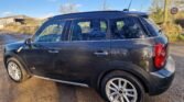 A black MINI Cooper Countryman is parked on a rural, sunlit dirt road. The car has four doors and silver alloy wheels. In the background, there is a wooden structure and greenery. A 30 mph speed limit sign is visible on the right side of the road, next to a stone wall. The sky is clear and blue.