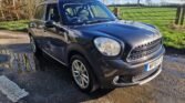 A black MINI Countryman parked on a paved area with puddles. Sunlight enhances its shine, reflecting off its well-polished surface. Surrounding environment includes a grassy field, a wooden fence, and trees. The sky is blue with a few clouds. The car's license plate reads "WF15 F".