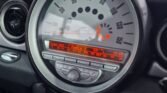 A close-up photo of a Mini Cooper's dashboard is shown. The speedometer, centrally located, dominates the image with a circular design displaying speeds in kilometers per hour. Below it, an illuminated digital screen shows radio station details, current time, and external temperature. Car controls are visible beneath the display of this automatic Mini 2007.