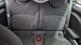 The image depicts the rear seat of a Mini 2007 with black fabric upholstery. The seat, showing slight signs of wear and discoloration, has dual headrests and is equipped with seatbelt buckles. The car door is visible on the right side of the image, emphasizing its compact, automatic design.