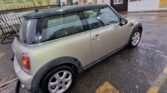 A silver 2007 Mini Cooper is parked outdoors in a car wash facility. The car has a black roof and black trim. The ground is wet, suggesting recent washing. The facility has a partially transparent roof, yellow pricing board, and a sign that reads "Hand Car Wash and Valeting." Various equipment is nearby.