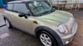 A silver Mini 2007 with a black roof is parked at a car wash facility under a blue-tinted canopy. The ground is wet, indicating recent rain or washing. A price and service menu for the automatic wash is visible on the brick wall in the background, along with metal chairs and a fenced area.