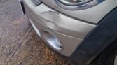 Close-up view of the front right corner of a silver 2007 Mini's bumper, showing minor damage including a noticeable dent and scuff marks. The headlight is intact, and droplets of water are visible on the car's surface, suggesting it was recently raining. The automatic car is parked on wet asphalt ground.