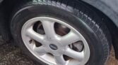 Close-up of a car's front wheel with a silver, five-spoke rim labeled "Mini" at the center. The tire is wet with water droplets, suggesting recent rain or washing. The surrounding area shows a wet, textured pavement and a partly visible body of what appears to be an automatic Mini 2007.