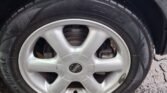 A close-up of a Mini 2007 Automatic's wheel and tire reveals a slightly deflated tire. The silver rim with five spokes glistens with water droplets. Wet conditions are indicated by the droplets on the vehicle's body and the wet pavement beneath the tire.