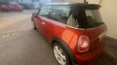 A 2013 Mini Cooper 1.6D with 128K miles, featuring a striking red exterior with a black roof and rear windows, is parked in a residential driveway. Viewed from the rear passenger side, white and brick houses and another red car are visible in the daylight background.