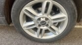 Close-up of a 2013 Mini Cooper's silver alloy wheel with the MINI logo at the center. The wheel has a five-double-spoke design and is fitted with a worn tire, resting on asphalt with a white parking space line. The car's fender is partially visible on the left, showcasing its sleek 1.6D model.