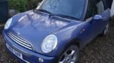 A blue 2005 Mini Cooper convertible is parked on a gravel driveway next to a green bush and a black shed. The car has a black soft top roof, circular headlights, and a license plate reading "NU55 SVR." Its exterior is slightly wet, indicating recent rain.