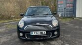 A black MINI Cooper with license plate LC60 MLK is parked on a cracked asphalt surface in front of a large, gray shipping container and next to a white building. Leafless trees and a hilly landscape are visible in the background under a partly cloudy sky.