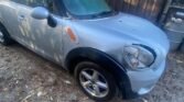 A silver Mini Cooper Countryman 2012 compact SUV with a slightly dusty windshield is parked on a dirt-covered surface next to a wooden building. The car's front left tire is slightly turned outward, and fallen leaves are scattered on the ground around the vehicle. A trash bin is partially visible in the background.