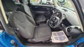 Interior of a MINI with black and grey cloth upholstery. The image shows the front seats, dashboard, and steering wheel from the passenger side door. The car has a manual transmission and various controls on the dashboard. A blue exterior and other parked vehicles are visible outside.