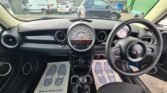 A MINI car's front interior view shows a steering wheel with the distinct Mini logo, a speedometer above the center console, and two footwell mats displaying blue footprints and text reading "Thank you for your custom." The car is parked in a lot with other vehicles in the background.