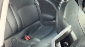 The image shows the interior of a Mini Cooper S, focusing on the backseat area. The seats are upholstered in black leather with visible seatbelts, including a bright red seatbelt buckle in the middle. The back of the front seat and part of the car door are also partially visible.