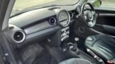 The interior of a small compact car, specifically a Mini Cooper S with a right-hand drive configuration. The dashboard features a central speedometer and air vents. The front seats are black leather, and the flooring has rubber mats. The gear shift and handbrake are in the center console. Outside, a brick wall and pavement are visible. ULEZ exempt and for sale!