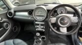 Interior view of a 3-door hatch, specifically a Mini Cooper S, showing the dashboard, steering wheel, and front seats. The dashboard features a circular speedometer and other gauges. The steering wheel, with a badge in the center, is on the right side. The gear shift and handbrake are visible between the seats.