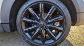 Close-up of a MINI Cooper's black, multi-spoke alloy wheel. The tire surrounding the wheel features the brand name "Cinturato," with its tread pattern in clear view. The car's white body and black wheel arch are partially visible, resting on a ground covered with small pebbles and gravel.