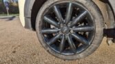 Close-up of a rear car wheel featuring a black alloy rim with multiple spokes and a MINI logo at the center. The weathered tire is mounted on a light-colored MINI vehicle, parked on a paved surface with a road sign and trees visible in the background. Lighting suggests it is daytime.