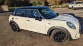 A white MINI Cooper with a black roof is parked in an outdoor parking lot. The car boasts black alloy wheels and is positioned at a slight angle, showcasing its side and part of the front. Several other vehicles and trees are visible in the background under a clear sky.