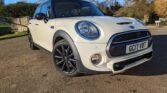 A cream-colored MINI Cooper is parked on a paved surface outdoors under a clear blue sky. The car features black alloy wheels, a black roof, and black side mirrors. Trees and foliage are visible in the background. The vehicle's license plate reads "GC17 VBT.