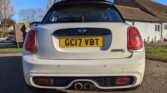 A white MINI Cooper S with the license plate "GC17 VBT" is parked on a paved area. This rear view shows its distinctive taillights and dual exhausts. A row of houses with sloped roofs and trees are visible in the background under a clear blue sky.