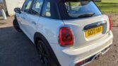 Side and rear view of a white MINI Cooper with a black roof parked on a paved surface. The license plate reads "GE17 VBT." The car features distinctive rounded tail lights and a sporty design. In the background, there is a white building, a green lawn, and trees under a clear sky.