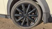 Close-up image of a car's black wheel rim with the manufacturer's logo "MINI" at the center. The vehicle's beige-colored body and part of the wheel arch are visible. The MINI tire is mounted on an asphalt ground under sunlight, casting shadows on the surface.