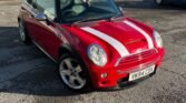 A red 2004 Mini Cooper S with white racing stripes is parked on a paved area in front of a metal building. The R53 model features a white roof, chrome side mirrors, and a UK license plate reading "YK54 CDX." The sun casts shadows on the ground under the clear sky. Trees and another vehicle are visible in the background.