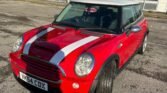 A red 2004 Mini Cooper R53 with white racing stripes is parked on a cracked asphalt surface in front of a blue and grey industrial building. The vehicle's license plate reads "YK54 CDX." The sun casts long shadows, and the sky is clear with a few clouds. Trees and greenery are visible in the background.