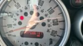 A close-up of a Mini Cooper S speedometer with a maximum speed reading of 160 mph. The digital odometer below the speedometer shows 35,086 miles. The image also reflects a partially visible hand and phone taking the photo. The car's dashboard and hazard lights button are partially visible on the right.