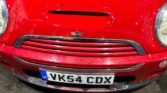 Close-up of the front of a red 2004 Mini Cooper S (R53) with a slightly dented grille and license plate "VK54 CDX." The car has visible water droplets, likely from recent rain, and is parked on a wet concrete surface. The background includes blurred objects and reflections.