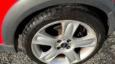 Close-up of the rear wheel of a red 2004 Mini Cooper S (R53), featuring a five-spoke alloy rim and a black tire. The wheel arch is framed with black trim. The car is parked on a rough, grey textured surface with small pebbles and scattered leaves. Sunlight casts shadows, adding depth to the image.