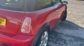 A red 2004 Mini Cooper S (R53) is parked in a lot with a slightly curved road to its right. The car's rear and left side are visible, including the license plate. The parking lot is bordered by a grassy area and industrial buildings are visible in the background under a clear sky.