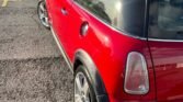 A red 2004 Mini Cooper S with a black roof and tinted windows is parked on a sunlit asphalt area. The car's rear quarter-panel and distinctive taillight are visible, and the scene includes a grassy area with a small staircase and a wooden fence in the background.