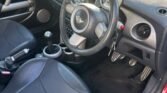 Interior of a 2004 Mini Cooper S featuring black leather seats, a black leather-wrapped steering wheel with the R53 car emblem, a manual gear shift, and various dials and controls on the dashboard. The car has a modern, clean design with visible foot pedals, and the door is open, letting in sunlight.