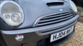 Close-up of the front of a gray MINI Cooper car, showing headlights, grille, and a license plate with the registration "HJ04 NHT." The car is surrounded by some gravel and trimmed hedges are visible in the background. The focus is on the MINI's details, highlighting its sleek design.