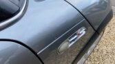 Close-up image of a grey MINI car's side view, focusing on the fender and a section of the door. The image highlights the S emblem located near the top of the fender, indicating the model's specification. The background shows a mix of gravel ground and a bit of the road.