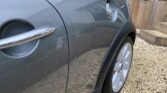 Close-up of a gray MINI's side, focusing on the rear wheel and door area. The car has a sleek metallic finish with a curved handle and chrome side detail. The shiny surface reflects its surroundings. The MINI is parked on a gravel driveway near wooden steps and a fenced garden area in the background.