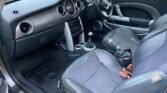 Interior of a MINI Cooper featuring black leather seats, a manual gear stick, and a dashboard with circular air vents and speedometer. The steering wheel proudly bears the MINI logo. Metal-patterned mats line the floor, and the handbrake is clearly visible beside the driver's seat.