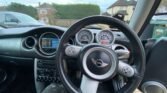 Wide-angle view of a MINI vehicle's interior focusing on the steering wheel and dashboard. The wheel displays the MINI logo and multiple controls. The dashboard includes various gauges and a digital display. Houses are visible through the windshield. The car’s gear shift and part of the driver's leg are also visible.