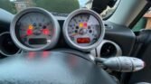 A car dashboard displays illuminated warning lights, including the check engine and low fuel indicators. The odometer reads 10150. Taken from the driver’s perspective of a MINI, the image shows the steering wheel in the foreground with houses and greenery visible in the background.