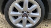 Close-up of a car wheel with a silver alloy rim designed with six spokes and a central piece that features a red "S" logo. The tire has visible text along the sidewall and is placed on a gravel surface. The MINI car body is partially visible on the upper edge of the frame.