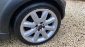 A close-up view of a MINI's front wheel featuring a silver alloy rim with a distinctive logo at the center. The tire is mounted and appears to be in good condition. The car's body is partially visible, and the ground is covered with small, light-colored gravel. A wooden fence is seen in the background.