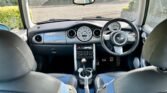 The image shows the interior of a 2004 Mini Cooper S, viewed from the back seat. The car boasts a sleek dashboard with a central speedometer, black leather seats with blue fabric inserts, a gear stick, and a three-spoke steering wheel. The view through the front windshield reveals a suburban street outside.