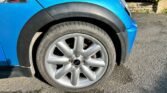 Close-up of the front left tire of a blue Mini Cooper S, specifically an R53 model with the Chilli Pack. The car's body features a black wheel arch trim, and the tire is mounted on a five-spoke alloy rim. The car is on a paved surface, with part of a stone wall and hedge visible in the background.