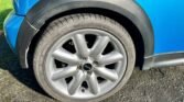 Close-up view of a 2004 Mini Cooper S wheel on a sunny day. The wheel features a silver rim with a black Mini logo in the center and is fitted with a Goodyear tire. The car, adorned in metallic blue, boasts smooth curves and black trim around the wheel well, characteristic of the R53 Chilli Pack.