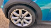 Close-up of a Mini Cooper S's front left wheel and part of the body. The rim is silver with five thick, rounded spokes, and the tire is black with a visible tread pattern. The turquoise blue body, featuring a Chilli Pack, has black trim around the wheel arch and an "S" badge above it. Parked on rough asphalt.